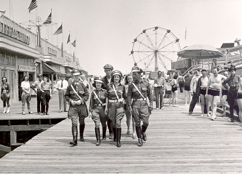 Historic image of motorcycle riders marching along a boardwalk with carnival attractions behind