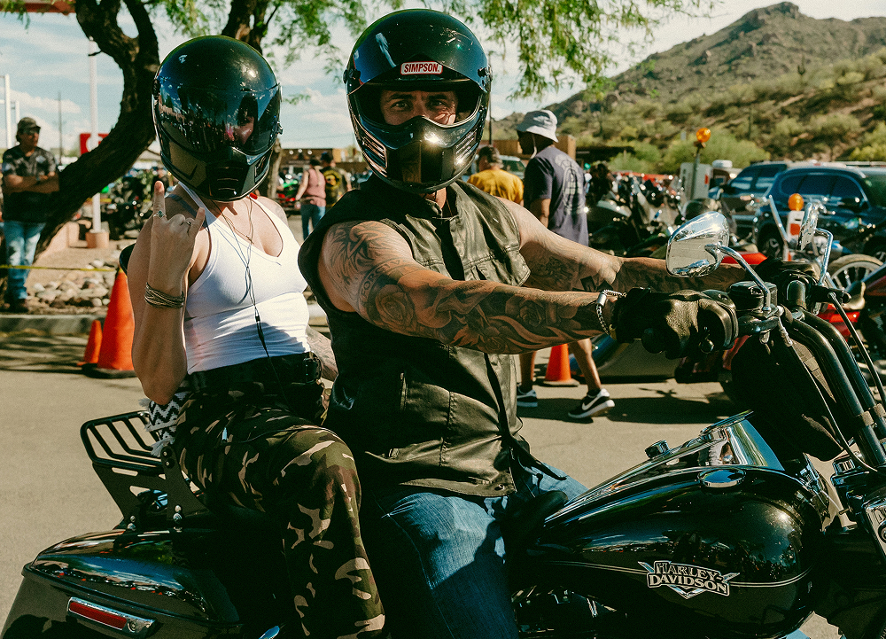 Two riders on a motorcycle parked at an event area, wearing helmets and gesturing towards the camera.