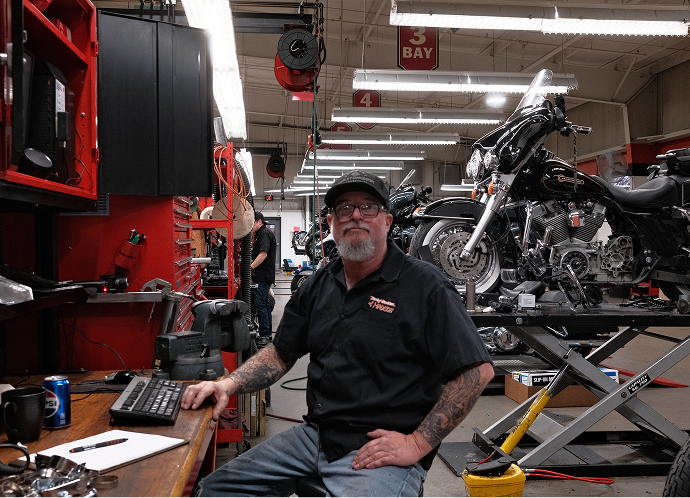 Harley‑Davidson technician seated at a workstation inside a service bay, with motorcycles on lifts and tools nearby