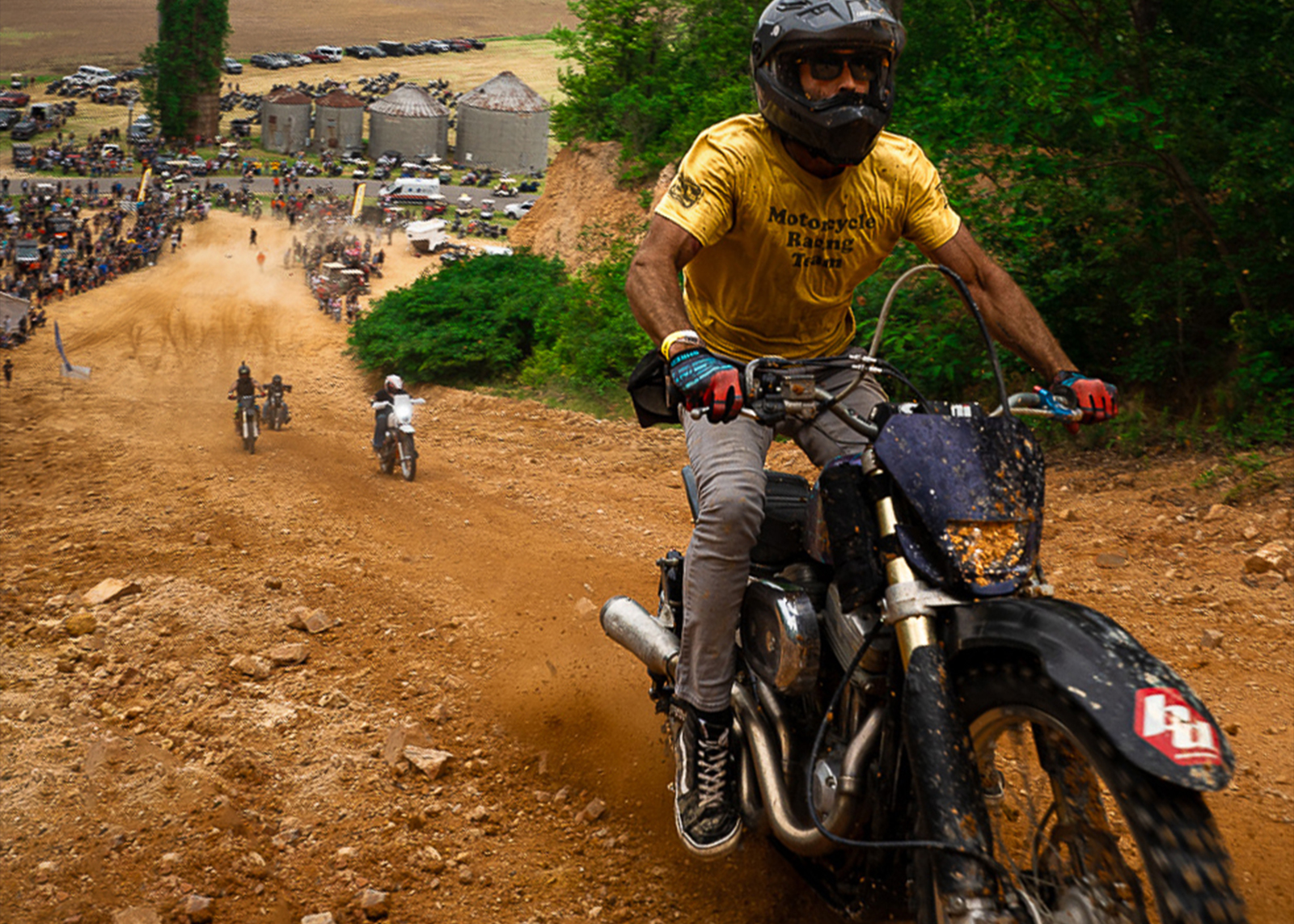 Four riders head up a dirt hill on their bikes with a large crowd in the background.