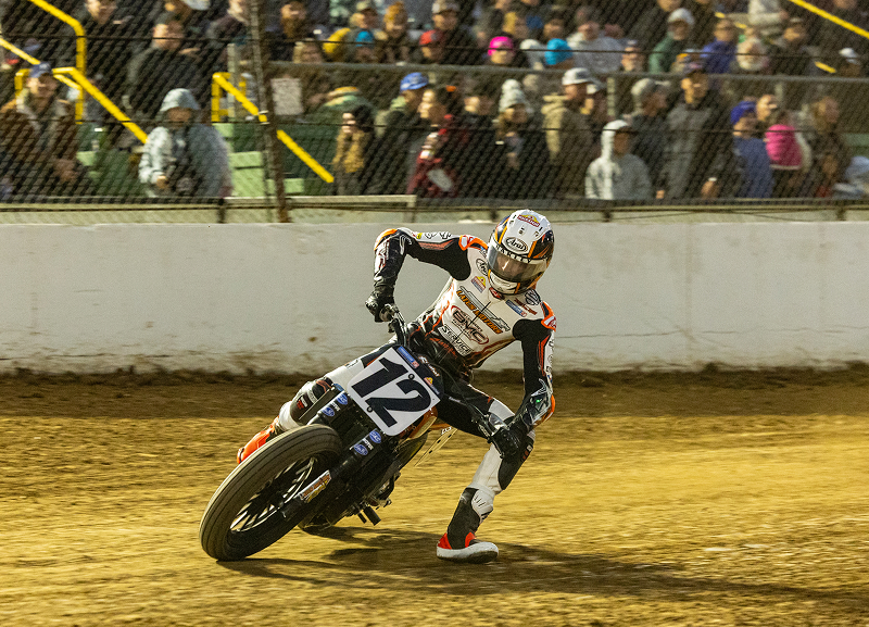 Motorcycle #12 slides through a dirt-track corner during a flat track race with an audience watching from behind