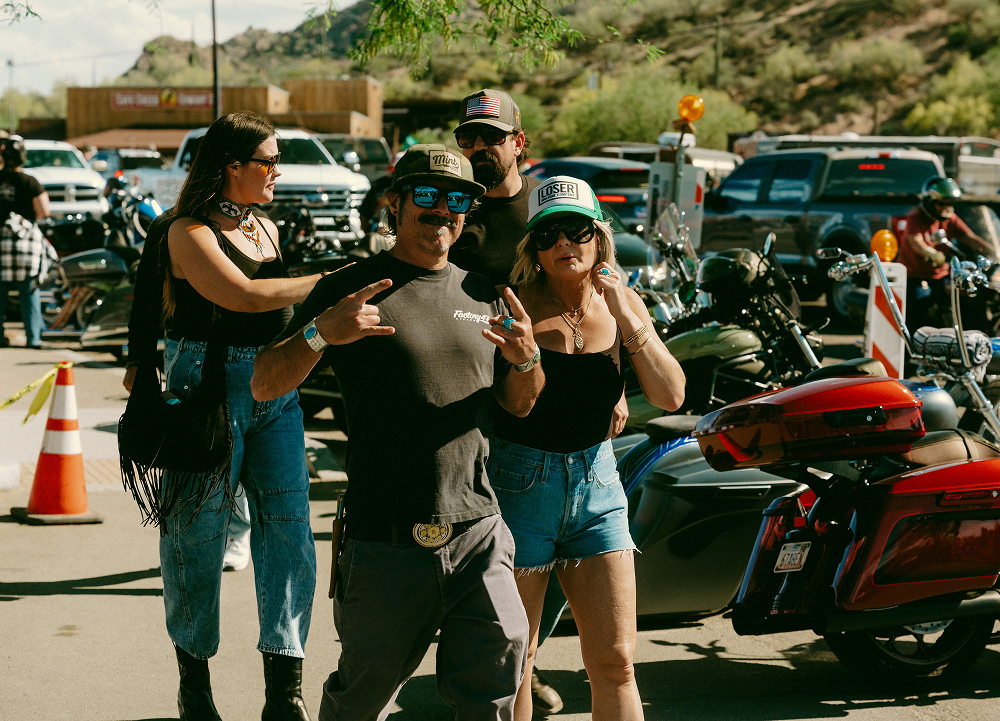 Four people walking near parked motorcycles at a crowded outdoor bike event with hills and trucks in background.