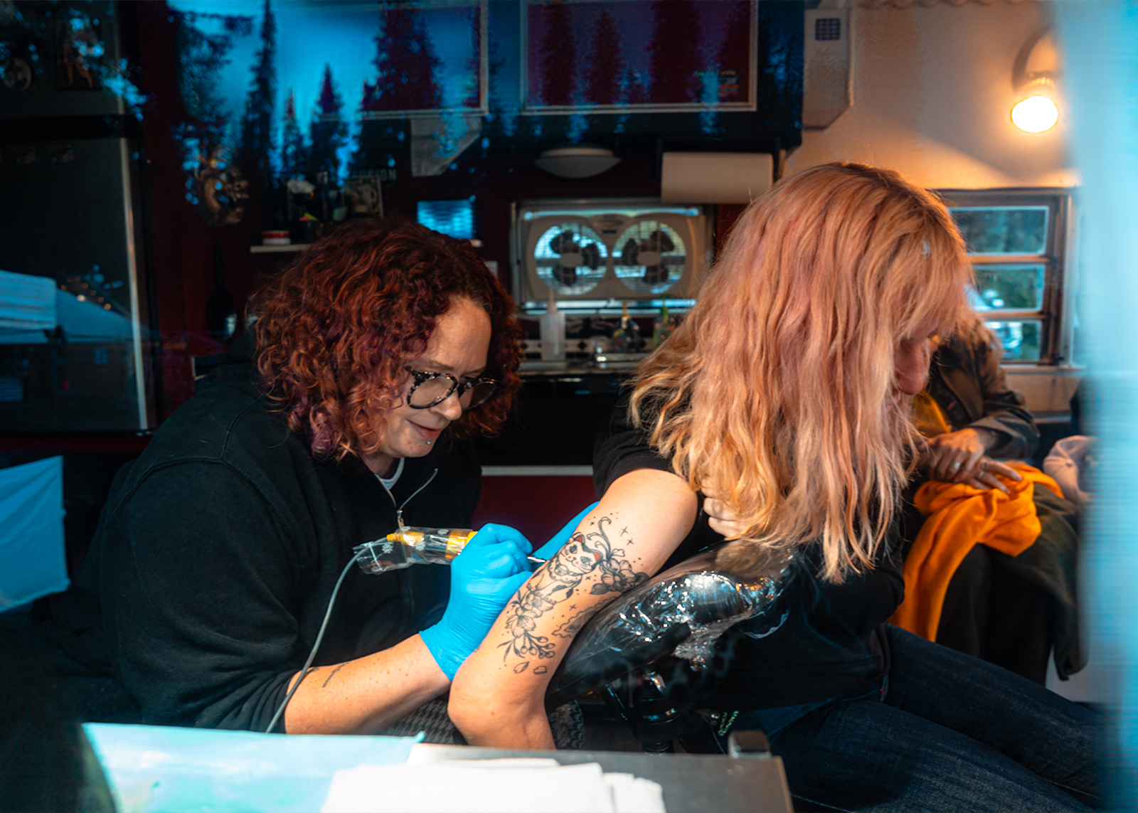 A smiling motorcyclist in a purple helmet and rain gear sits on a bike among trees and parked motorcycles.