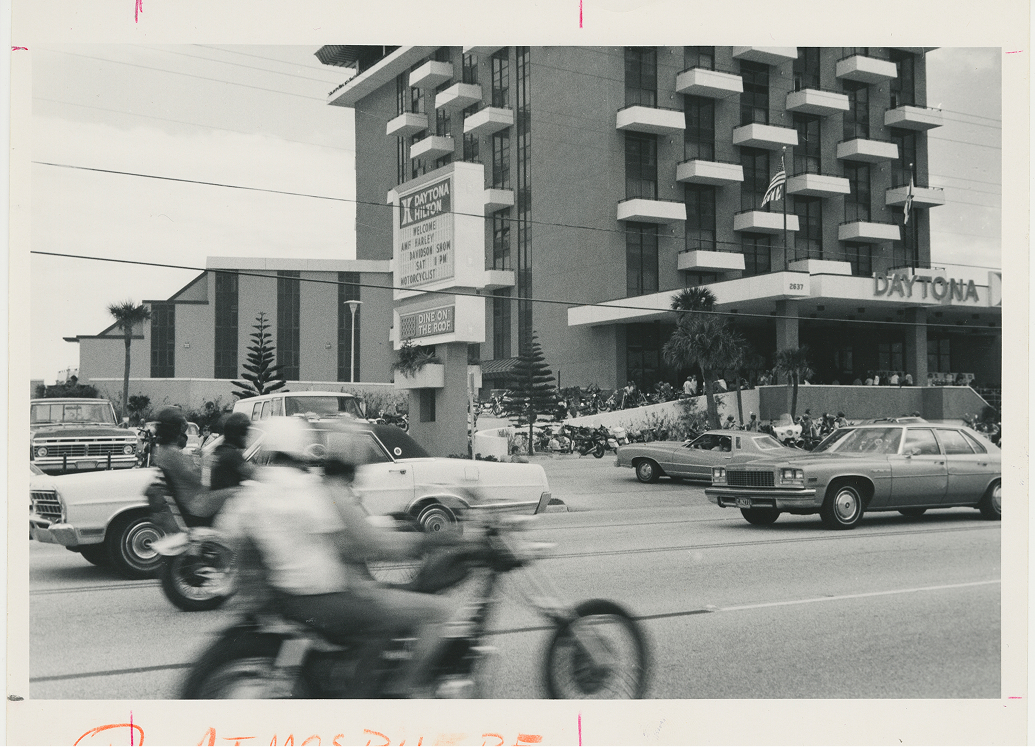 Black‑and‑white photo of motorcycles and cars moving past the Daytona Hilton with a crowd gathered near the entrance