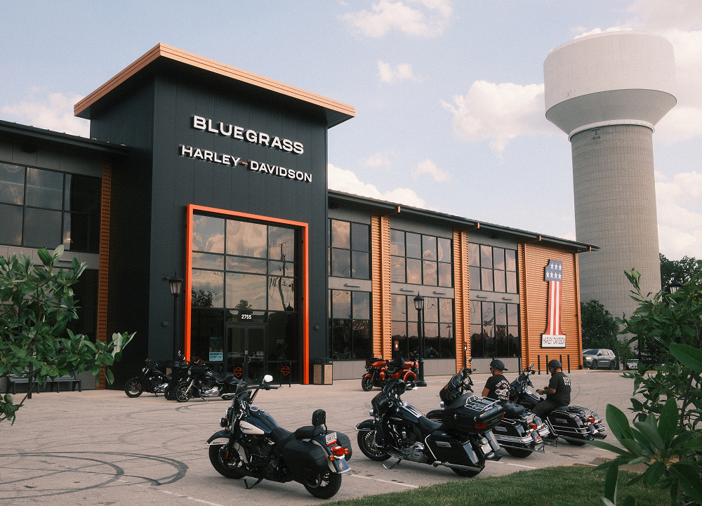 Exterior view of Bluegrass Harley-Davidson dealership with black and orange facade, large windows, and water tower nearby