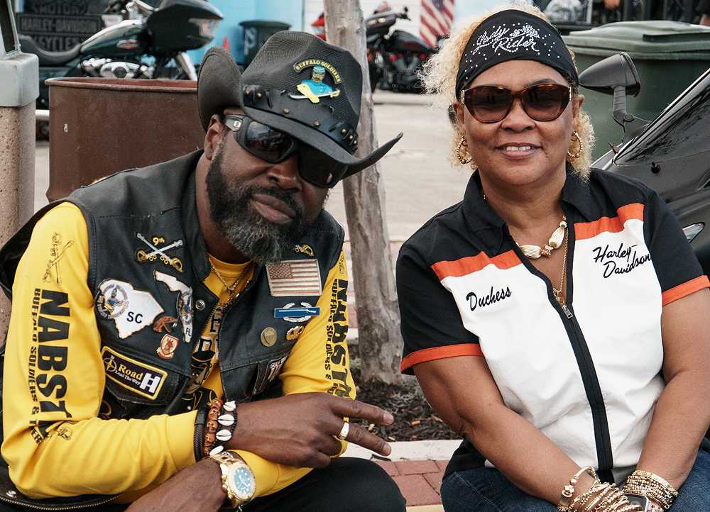 Two people sit side by side wearing motorcycle vests, with parked bikes and event activity behind them