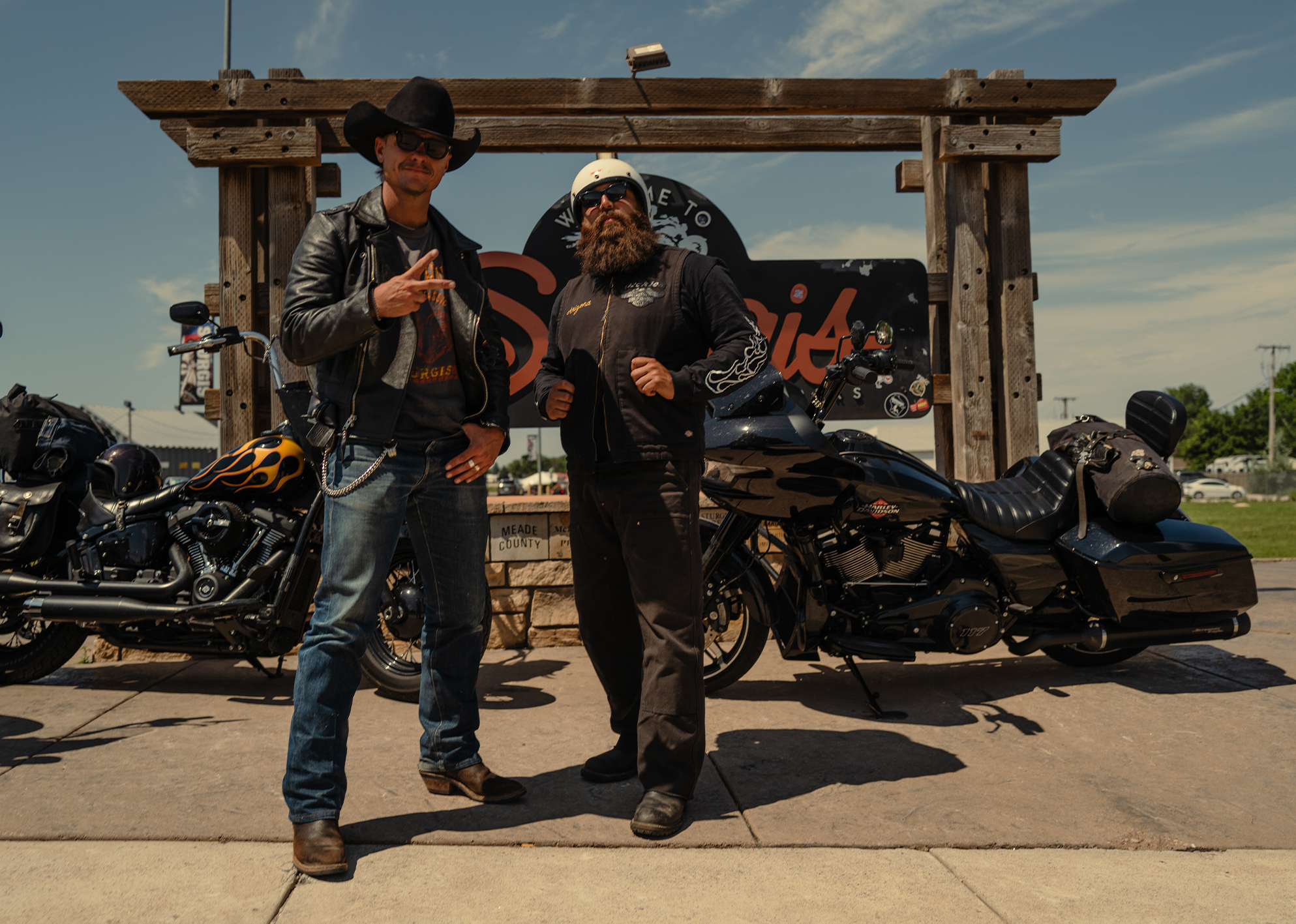 Two bikers pose in front of the Sturgis sign with their Harley-Davidson motorcycles.
