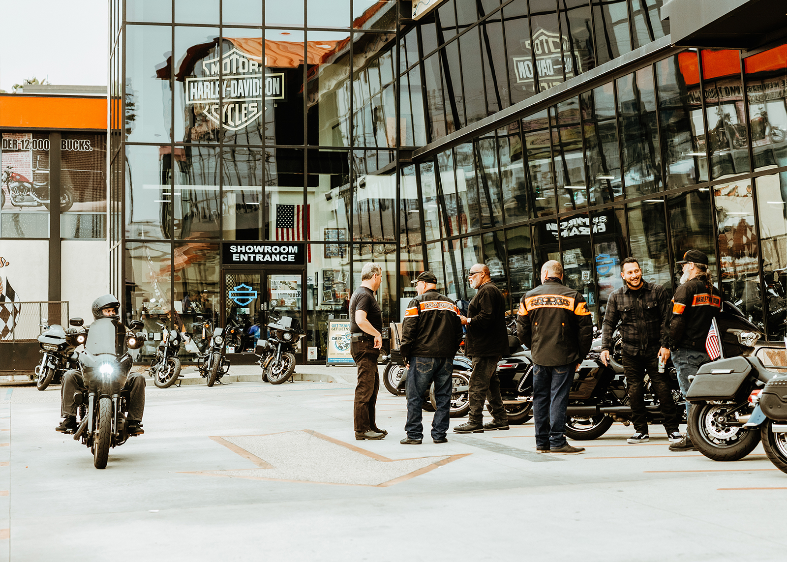 A group of riders outside Bartel’s Harley-Davidson watch another rider pass by in the parking lot.