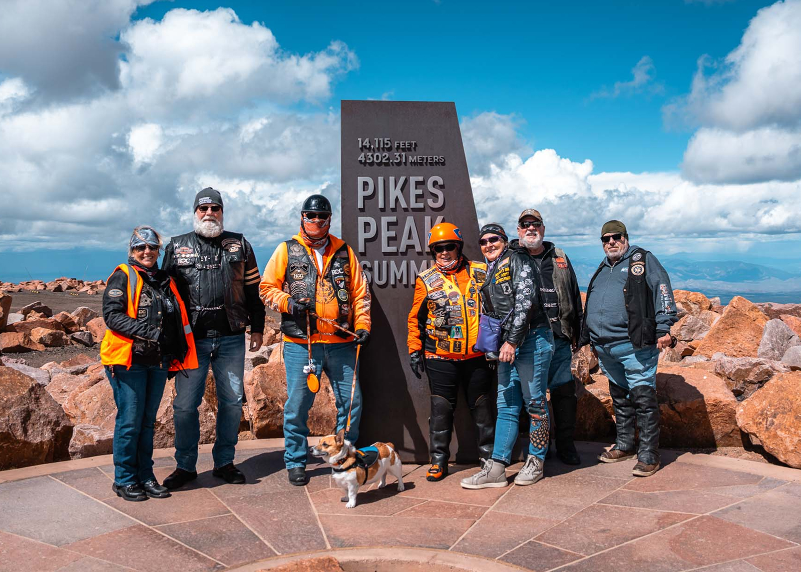 Group of bikers and a Corgi posing at the Pikes Peak Summit marker surrounded by rocks under a bright sky