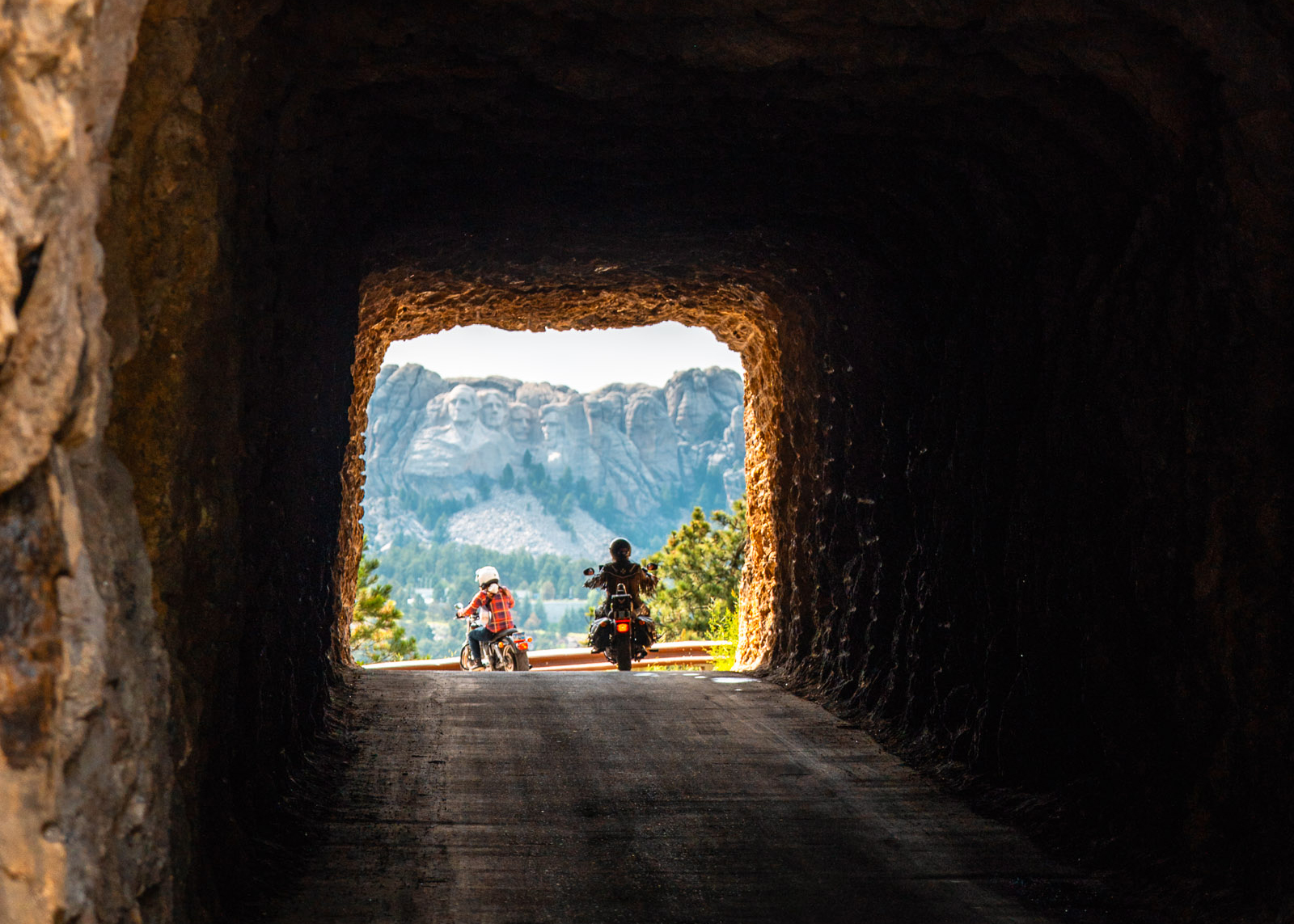Two motorcyclists framed by a tunnel exit with Mount Rushmore visible in the distance through the opening.
