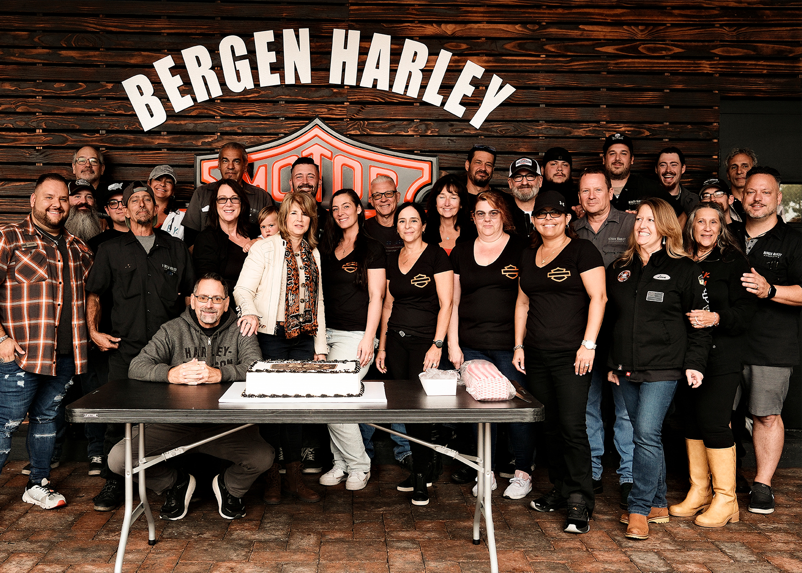 The Bergen H-D family is gathered under a Bergen Harley sign, smiling behind a table with a cake.