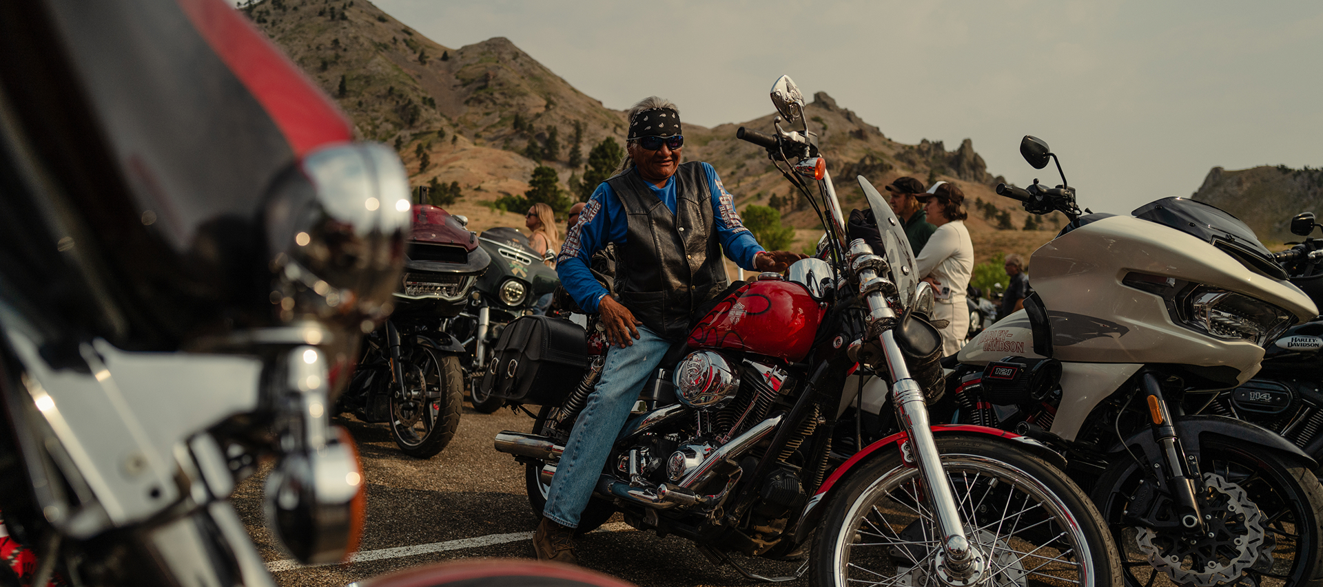 Motorcyclist on a custom red Harley-Davidson parked among riders and bikes in a mountain gathering area