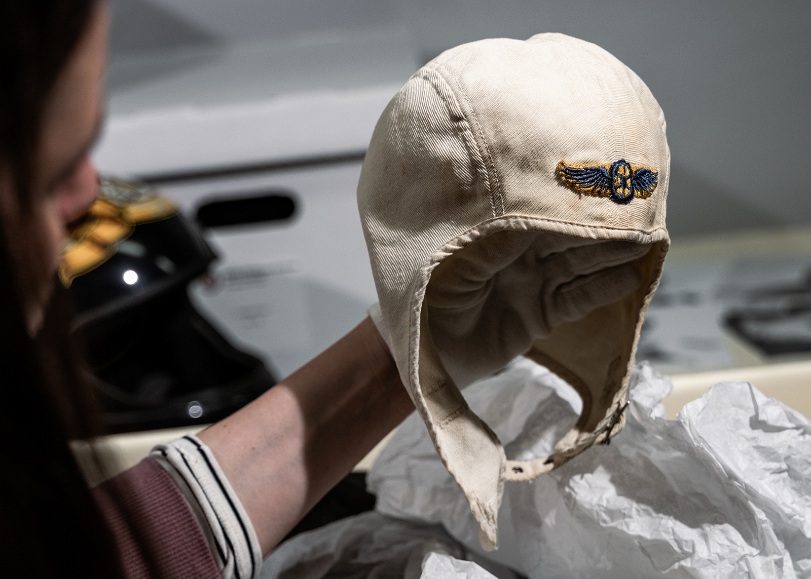 A museum archivist holds a vintage white canvas motorcycle helmet with a winged wheel emblem embroidered on the front.